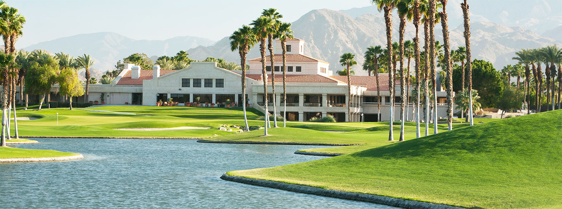 A scenic view of a golf course with palm trees, a lake, and a clubhouse against a mountain backdrop.