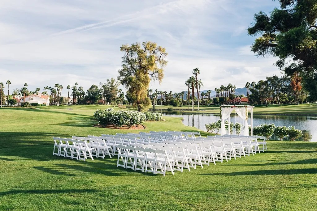 Outdoor wedding setup with white chairs arranged near a lake, decorated arch, and palm trees in the background.